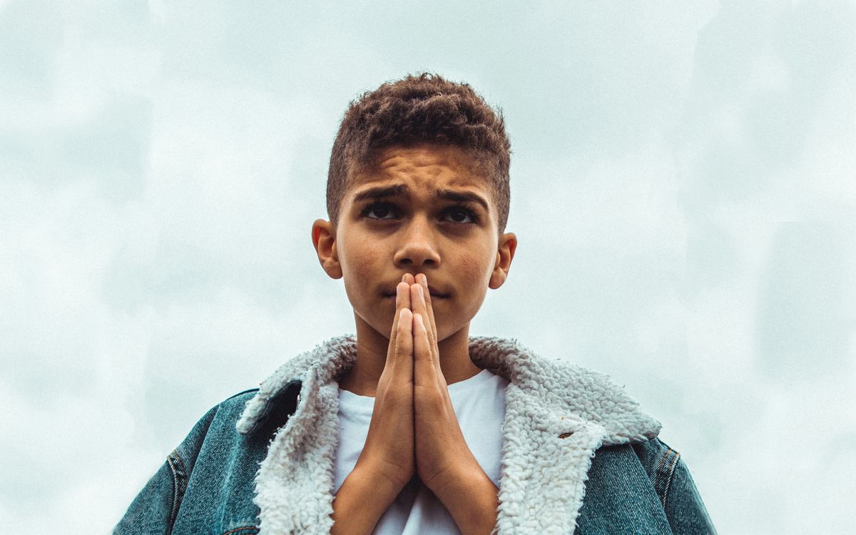 A boy in a denim jacket looking up, hands joined.