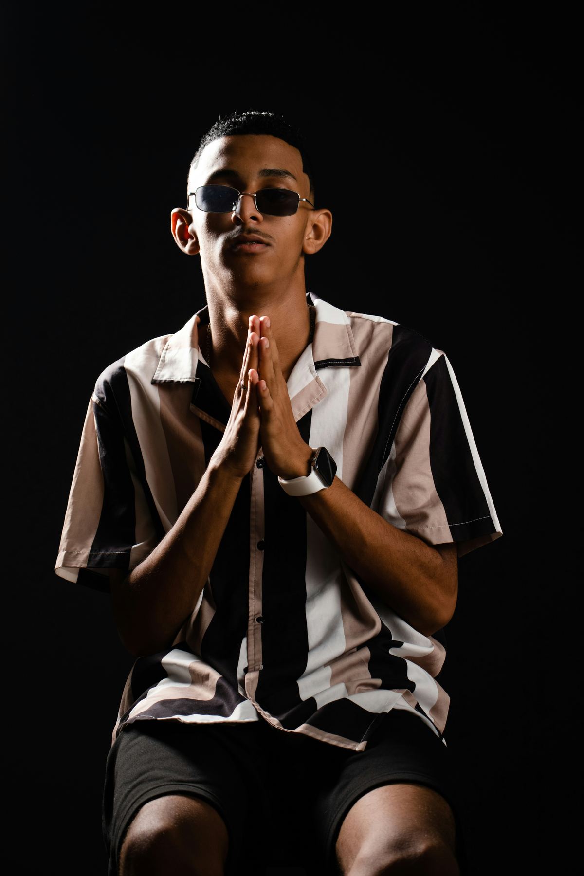 A young man sitting against a dark background, sunglasses on, hands joined in prayer.